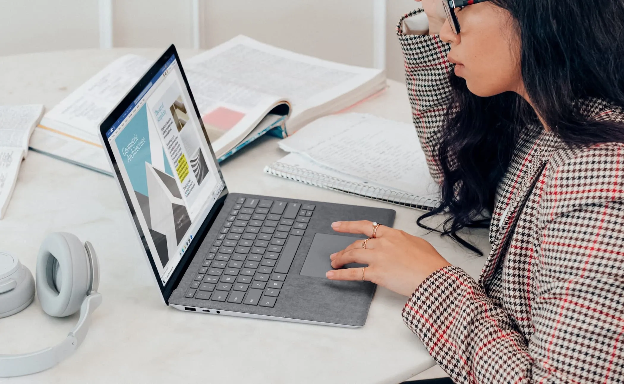 Frau am Laptop bearbeitet ein Layout, umgeben von aufgeschlagenen Büchern, Notizen und Kopfhörern.