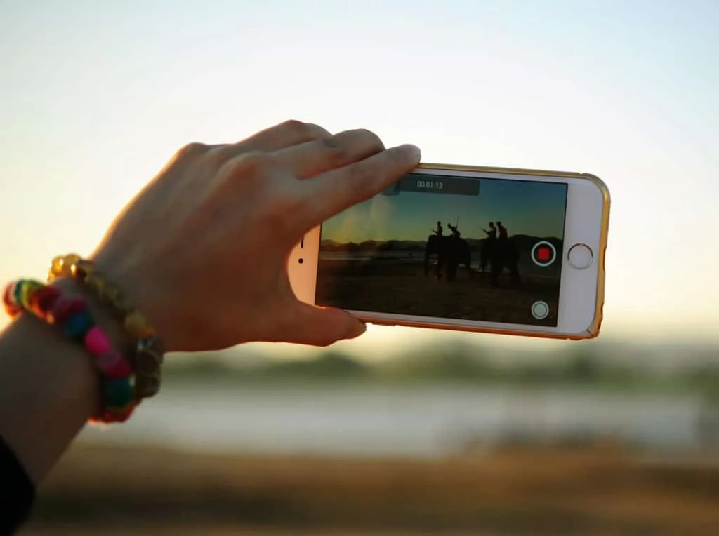Una mano filma con lo smartphone al crepuscolo; sullo schermo persone in sella a elefanti e il timer di registrazione