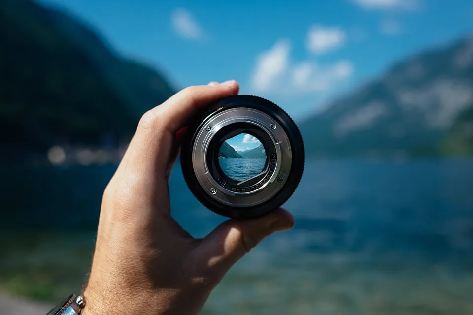 Hand hält ein Kameraobjektiv vor eine Berglandschaft; durch das Glas wird der See scharf sichtbar.