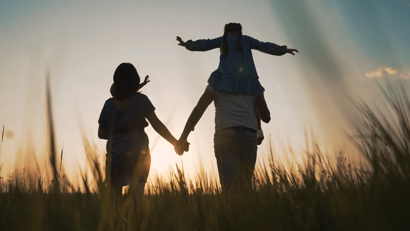Familie im Gegenlicht: Der Vater trägt die Tochter auf den Schultern, daneben geht die Mutter durch hohes Gras