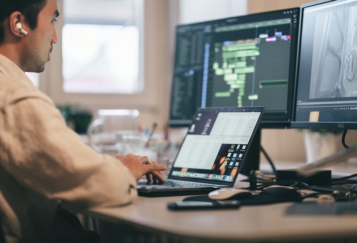 Ein Mann arbeitet am Laptop vor zwei Bildschirmen mit Videoschnitt.